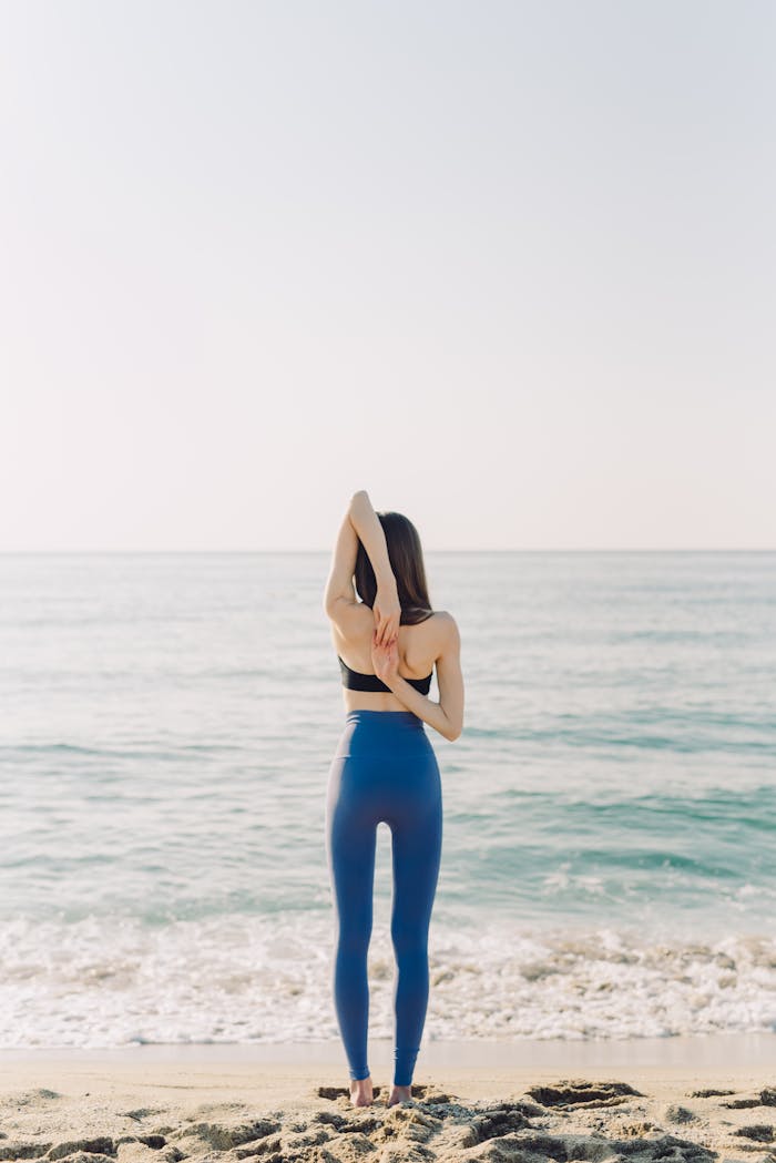 Rear view of a woman practicing yoga on a serene beach under a bright sky.